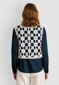 Ručni šal s cvjetnim uzorcima u krem i tamnoplavoj boji, s valovitim rubom i otvorenim leđima, nosi se preko tamnoplave košulje dugih rukava.