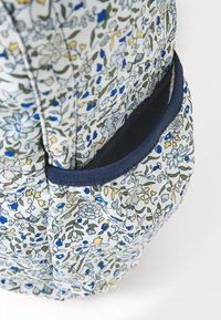Rucksack aus floral gemustertem Stoff mit einer Vielzahl von blauen, gelben und grünen Blumen auf weißem Hintergrund und einer dunkelblauen Taschenkante.