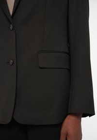 Tumehall stiililine pintsak, mis on valmistatud tekstuursetest kangast. Omab kahte nuppu, ühte taskut vasakul ja struktureeritud õlavarre osasid, millel on sile viimistlus.