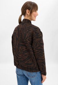 Woman with brown hair in a ponytail wearing gold hoop earrings, a black and brown patterned jacket, and blue jeans, smiling facing sideways.