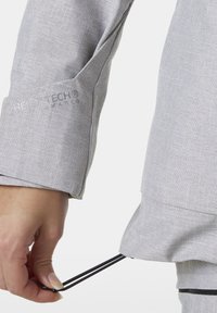 Světle šedý, texturovaný detail bundy ukazuje ruku, která upravuje elastický stahovací pás na spodním lemu, s tištěným logem na rukávu.