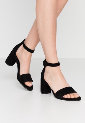 Zwarte sandalen van suède met blokhak, een enkelbandje en open teen, voorzien van een brede voorband en een gestructureerde afwerking.