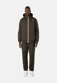 Felpa con cappuccio verde scuro, con zip, coulisse e branding bianco, abbinata a pantaloni jogger coordinati e sneakers bianche. Tessuto dalla texture liscia.