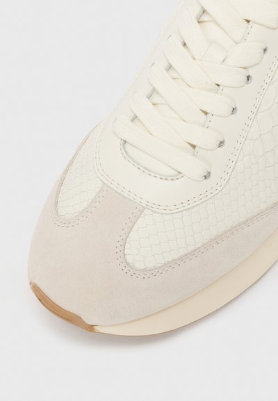 Suede, tekstuuriga nahast paneelidega off-white spordijalatsite nina lähivõte, millel on kreemjad seotud paelad valgel taustal.