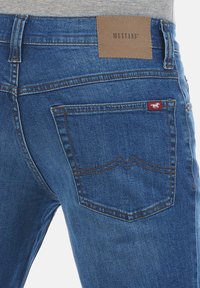 Jeans de mezclilla azul con un parche de cuero marrón etiquetado como "MUSTANG", bolsillo trasero visible con una etiqueta de logo roja y costuras en contraste.