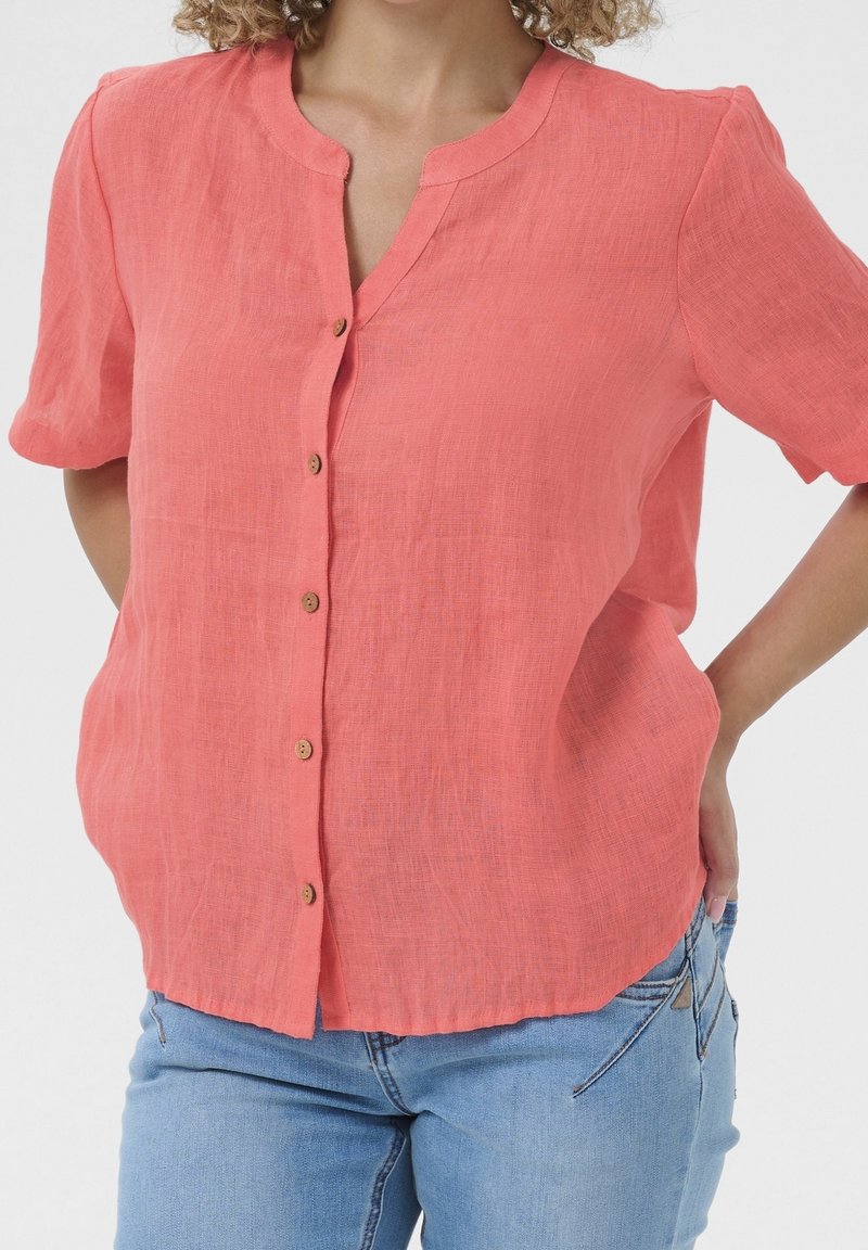 Femme portant un chemisier corail à manches courtes boutonné, associé à un jean bleu clair, debout les mains derrière le dos.
