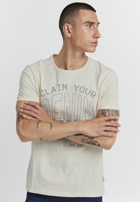 Camiseta color crema con escote redondeado, que presenta el gráfico "RECLAMA TU DENIM" en marrón oscuro, mangas cortas y corte casual.
