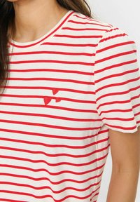 Primer plano de una persona que lleva una camisa blanca con rayas rojas horizontales y tres pequeños corazones rojos bordados en el pecho.
