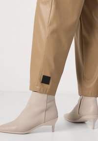 Beige nepleren broek met een zwarte logopatch aan de zoom, gecombineerd met beige puntige enkellaarsjes met lage hakken en een gladde textuur.