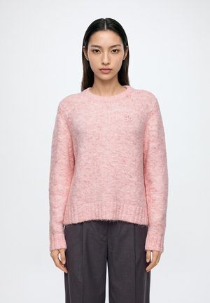 Una mujer luce un jersey de punto rosa pálido con cuello redondo y unos pantalones oscuros sobre un fondo liso y claro.