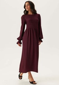 Robe longue bordeaux avec un corsage smocké, des manches évasées et une jupe fluide. Tissu léger avec un design texturé et sans embellissements.