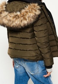 Chaqueta acolchada color verde oliva que presenta una capucha con piel sintética desmontable, diseño acolchado y puños elásticos, combinada con vaqueros de mezclilla azul claro.