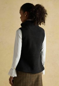 Čierna fleecová vesta s vysokým golierom, s hladkou textúrou a prispôsobeným strihom, nosená cez biele tričko s volánovými manžetami.