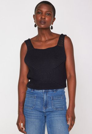 Eine Frau trägt ein ärmelloses schwarzes gehäkeltes Oberteil mit breiten Trägern und eine hellblaue hochgeschnittene Jeans, steht vor einem schlichten weißen Hintergrund.