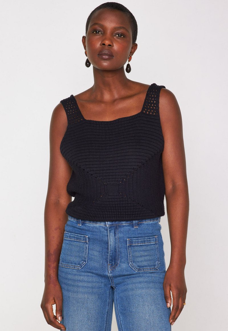 Donna che indossa un top nero in cotone all'uncinetto senza maniche con spalline larghe e jeans a vita alta blu, in piedi contro uno sfondo bianco uniforme.