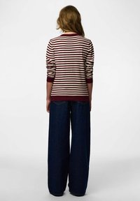 Tumši sarkans un krēmkrāsas svītrains džemperis ar ribām piedurknēs un apakšmalā, kombinēts ar plašā piegriezumā tumšām džinsa biksēm. Modelis redzams no aizmugures.