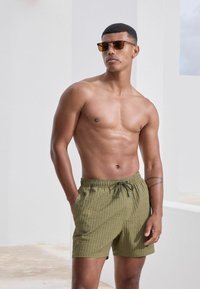 Olivengrüne Badehose mit strukturiertem Muster, elastischem Bund und Kordelzug. Getragen von einem Modell mit Sonnenbrille, das einen lässigen Look zeigt.
