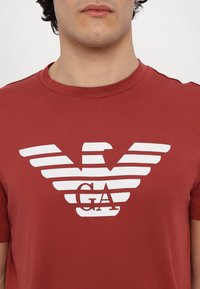 Czerwona bawełniana koszulka z krótkimi rękawami, z białym logo orła w paski i napisem "GA" na środku, wykonana z gładkiej tkaniny.