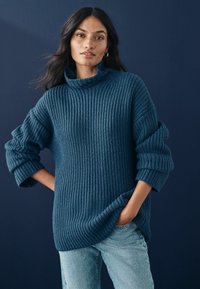 Pulover tricotat de culoare albastră închisă, cu guler înalt, croială lejeră și textură ribborată. Mâneci răsfoite și slituri laterale contribuie la designul său relaxat.