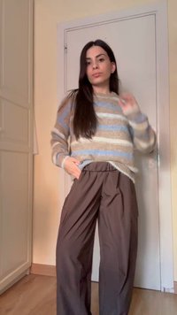 Pikkade tumepruunide juustega naine, kes kannab beežide, siniste ja valgete triipudega kampsunit ning lahtisi pruune pükse, seisab siseruumis.