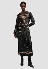 Vestido longo preto de lantejoulas com bordados dourados, apresentando padrões de estrelas e flores, mangas longas e decote redondo. Combinado com botas pretas.
