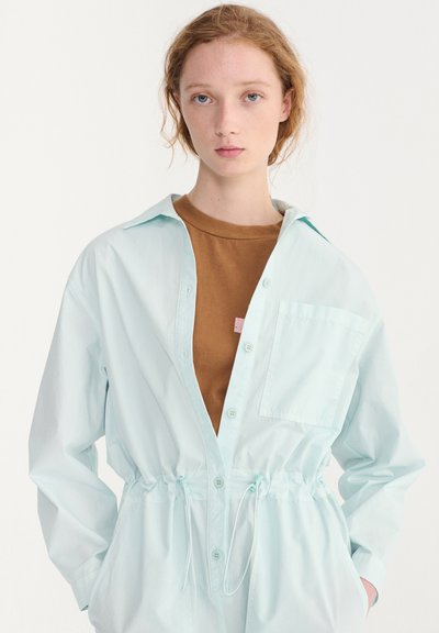 Jeune femme aux cheveux roux bouclés portant une veste bleue claire boutonnée par-dessus une chemise marron, debout devant un fond blanc uni.