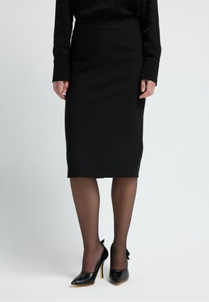 Zwarte kokerrok met een gladde textuur, gecombineerd met zwarte hoge hakken met een strikaccent aan de achterkant. Transparante zwarte tights maken de look compleet.
