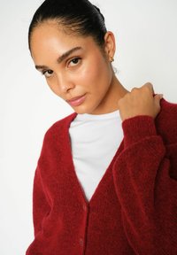 Cardigan tricotat roșu, cu decolteu în V, nasturi în față și material texturat. Purtați pe un tricou alb, cu mâneci ajustate.