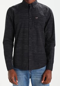 Uomo che indossa una camicia nera con bottoni e un piccolo logo rosso e bianco sulla tasca del petto, accompagnata da jeans blu.