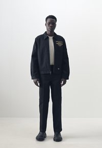 Black bomber jacket with logo, white crew neck shirt, black trousers, and black shoes. Model stands against a plain background.