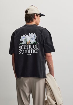 Mann iført svart T-skjorte med hvite blomster og teksten "sommers duft" på ryggen, beige bukser, krem og svart caps, holder en lett jakke.