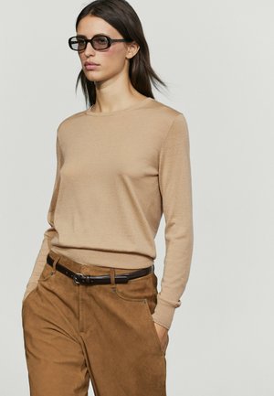 Femme aux cheveux foncés portant des lunettes de soleil noires, un pull beige à manches longues, un pantalon marron et une ceinture noire, les mains dans les poches.