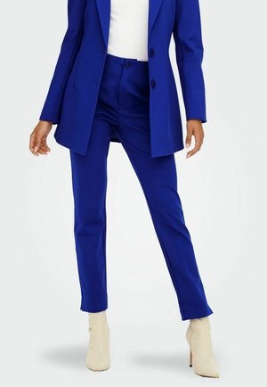 Femme portant un tailleur bleu roi avec un pantalon et un blazer sur mesure, associé à des bottines blanches et un haut blanc.