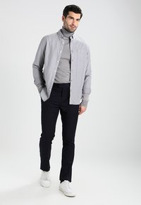Grå skjorte med knapper og et subtilt mønster, lagdelt over en lys grå høyhalset genser, parret med marineblå bukser og hvite joggesko.