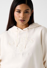 Roomkleurige hoodie met trekkoorden, voorzien van een opgehoogd "GUESS" logo en geborduurde tekst. Zachte stof en ontspannen pasvorm.