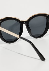 Occhiali da sole neri con finitura lucida, forma a occhio di gatto oversize e lenti scure. Accenti in tono oro sulla parte superiore e aste in marrone chiaro.