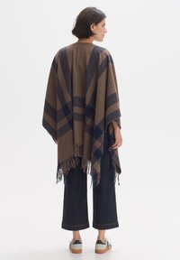 Wool poncho in brown with navy plaid pattern and fringed hem, loose fit, paired with dark cropped jeans and casual shoes.