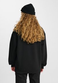 Persona con lunghi capelli biondi ricci, che indossa un berretto nero e un felpa nera larga, in piedi di spalle su uno sfondo neutro.