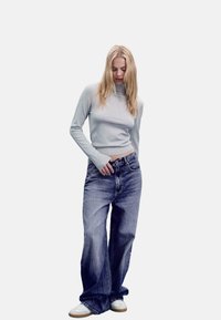 Hellgraues, tailliertes Langarmshirt kombiniert mit übergroßen blauen Jeans. Das Model trägt weiße Sneakers. Schlichte, weiche Texturen und lässiges Design.