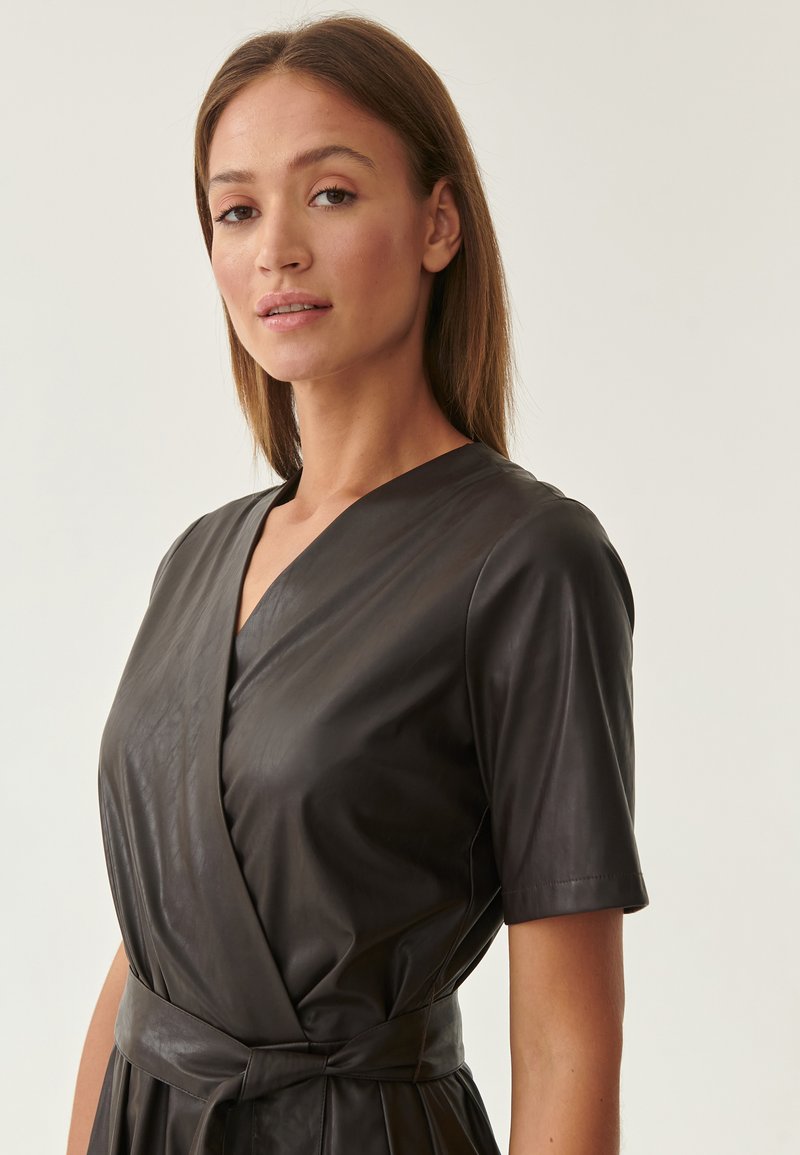 Giovane donna con capelli castani lisci che indossa un vestito avvolgente in pelle nera a maniche corte con una cintura legata su uno sfondo neutro.