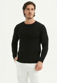 Un pull en tricot noir à col rond avec de longues manches, design ajusté et poignets côtelés, associé à un pantalon blanc. Texture lisse, sans motifs.