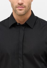Camicia nera con bottoni, caratterizzata da sottili righe bianche verticali, colletto strutturato e un leggero luccichio. Il tessuto appare liscio con una vestibilità su misura.