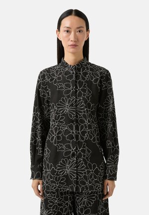 Zwarte blouse met wit bloemenpatroon, lange mouwen en een mandarinakraag. Met knopen aan de voorkant en zachte, gestructureerde stof.