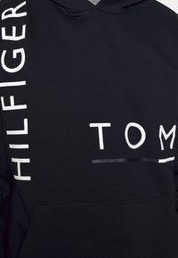 Black hoodie with white printed text "HILFIGER" and "TOM" on the front. Features a kangaroo pocket and a soft texture.