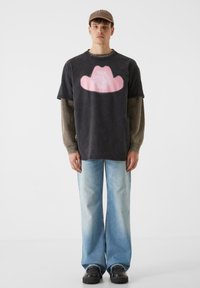 Melnas kokvilnas T-krekls ar lielu rozā kovboja cepuri, kas izcelta uz krūšu daļu, kopā ar gaiši ziliem izsistiem džinsiem un melnām kurpēm.