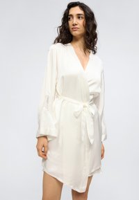 Γυναίκα με σγουρά σκούρα μαλλιά που φοράει λευκό σατεν φόρεμα τύπου wrap με μακριά μανίκια και ζώνη στη μέση, κοιτάζει προς τα πλάγια.