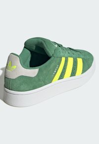 Zapatillas de ante verde con tres rayas amarillas, suela de goma blanca y acento en el talón gris. Presenta un empeine texturizado y detalles del logotipo.