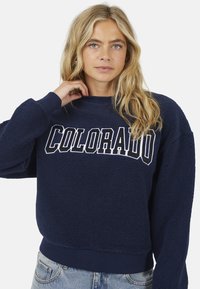 Donkerblauwe fleece sweatshirt met "COLORADO" tekst in het wit, geribbelde boorden en een cropped ontwerp. Zachte textuur en losse pasvorm.