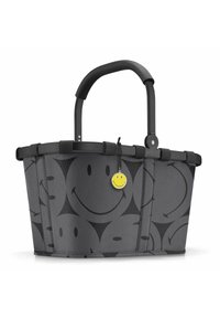 Cesta de compras de tela negra y gris con un patrón de caras sonrientes, estructura rígida y un mango de goma. Incluye una etiqueta amarilla con una sonrisa.