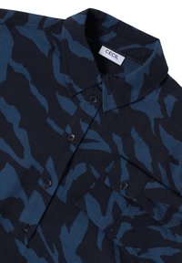 Camicia a maniche corte di colore blu scuro realizzata in tessuto testurizzato. Presenta un colletto, chiusura con bottoni e un motivo ondulato simile al camouflage in diverse sfumature di blu.